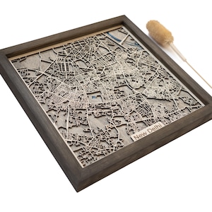 Puede incluir: Un mapa de madera enmarcado de Nueva Delhi, India, con intrincados detalles cortados con láser. El mapa está enmarcado en un marco de madera gris oscuro. Las calles y edificios de la ciudad están representados con precisión en un tono de madera clara. Un tallo de flor seca decorativo está en el fondo.