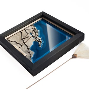 May include: A framed wooden map of the coast of  "Cabo Coco Cuba" with a blue resin inlay. The map is in a black frame and is sitting on a white surface.