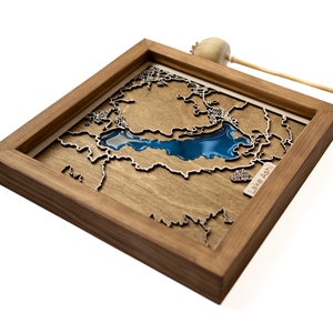 Puede incluir: Un mapa enmarcado de madera del lago Ashti con una incrustación de resina azul que representa el lago. El mapa está montado sobre un fondo de madera.