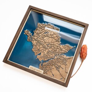 May include: A wooden map of the Isle of Anglesey, Wales, with a blue resin background. The map is framed in a dark wood frame and includes the text "Ynys Môn".