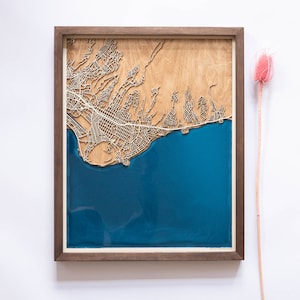 A wooden map of your favourite place enclosed in a wooden frame. It has 3D city streets and epoxy resin water. By adding a pin in a important place for you and an engraving you will make a personalized gift.