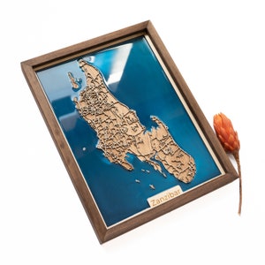 May include: A wooden map of Zanzibar, framed in a dark wood frame. The map is set in a blue resin, giving the appearance of water. The frame is on a white surface.