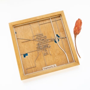 May include: A wooden map of Tremont, Illinois, in a light brown frame. The map is laser cut and shows the streets and waterways of the town. The map is mounted on a light brown background.