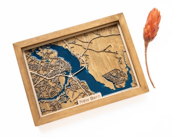 May include: A wooden map of New Bern, North Carolina, with a blue resin inlay representing the water. The map is framed in a natural wood frame.