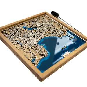 May include: A wooden framed map of Baku, Azerbaijan, with a blue resin inlay representing the Caspian Sea. The map is detailed and shows the city's streets and landmarks.