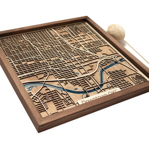 Puede incluir: Un mapa de madera del centro de OKC, enmarcado en un marco de madera oscura. El mapa está cortado con láser y presenta un río azul que atraviesa la ciudad.
