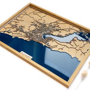 Puede incluir: Un mapa de madera de Dunedin, Nueva Zelanda, con una incrustación de resina azul que representa el océano. El mapa está enmarcado en un marco de madera natural.