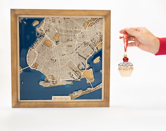 Pode incluir: Um mapa de Brooklyn, Nova Iorque, emoldurado em madeira com um esquema de cores azul e dourado. O mapa é um mapa em relevo 3D com o texto "Brooklyn" na parte inferior. Uma mão está segurando um enfeite de cupcake vermelho e branco.