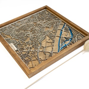 May include: A wooden map of Vienna, Austria, with a blue river and the city name "Vienna" in the lower left corner. The map is framed in a natural wood frame.