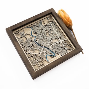 May include: A wooden framed map of Monroe, LA. The map is laser cut and features a blue river running through the city. The frame is dark brown.