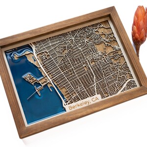 May include: A wooden framed map of Berkeley, CA with a blue resin inlay. The map is laser cut and shows the city streets and waterways.
