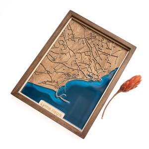 May include: A wooden map of Lyme Regis with a blue resin inlay representing the ocean. The map is framed in a dark wood frame.