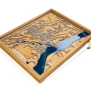 May include: Wooden framed map of Kingston, NY, with intricate laser-cut details. The map features a light brown land area, a blue water section, and a white label with the city name. The frame is a light brown color.