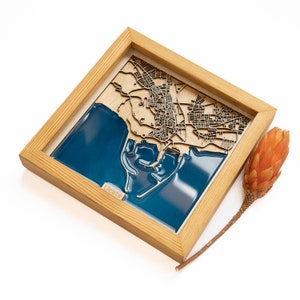 May include: A wooden framed map of Licata, Italy, with a blue resin inlay representing the water. The map is laser cut and features a detailed representation of the city streets.