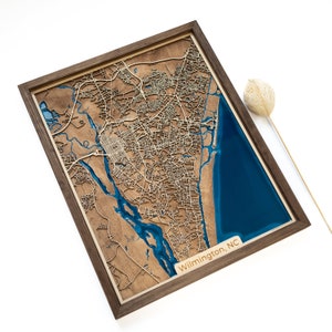 Op de afbeelding: Een houten kaart van Wilmington, North Carolina, met een blauwe harsinleg die het water voorstelt. De kaart is ingelijst in een donkerhouten lijst.