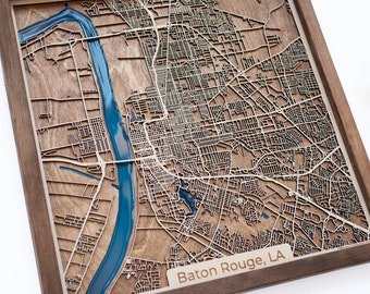 May include: A wooden map of Baton Rouge, Louisiana, with a blue river running through it. The map is framed in a dark wood frame.