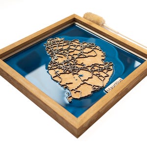 May include: A wooden map of Mauritius in a light wood frame with a blue resin background. The map is detailed with roads and cities. The word "Mauritius" is written in the bottom right corner of the frame.