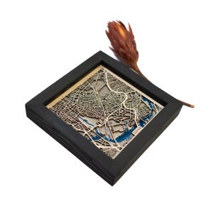 May include: A black wooden frame containing a laser-cut wooden map of a city. The map is in shades of brown and blue, with the city's waterways highlighted in blue. The frame is on a white surface. The text "02149" is visible in the bottom right corner of the map.