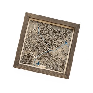 May include: A wooden map of Guelph, Ontario, in a brown frame. The map is laser cut and shows the city's streets and landmarks. The word "Guelph" is printed at the bottom of the map.