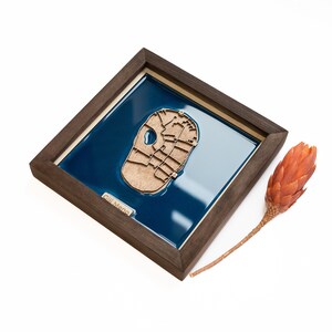 May include: A wooden map of Gill Meno, a small island, is framed in a dark wood frame. The map is set against a blue background. The frame is sitting on a white surface.
