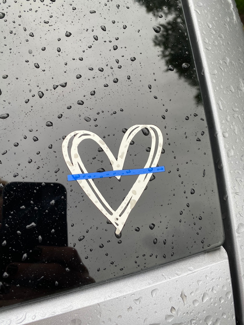 K&ouml;nnte beinhalten: Ein wei&szlig;er Herzaufkleber mit einem blauen Streifen in der Mitte, auf einem Autofenster mit Regentropfen.