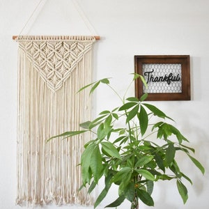 May include: A white macrame wall hanging with a geometric pattern, a potted green plant, and a wooden frame with the word "Thankful" on a white wall.