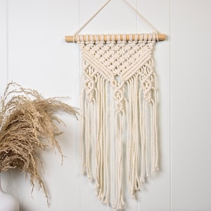 May include: A white macrame wall hanging with a geometric pattern. The hanging is made of cotton cord and has a wooden dowel at the top.
