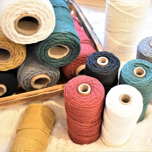 Puede incluir: Surtido de carretes de cuerda de algodón de colores en varios colores, incluyendo blanco, verde, negro, rojo y amarillo. Los carretes están dispuestos en una caja de madera y sobre una superficie blanca y esponjosa.