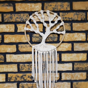 May include: A white macrame wall hanging in the shape of a tree with a wooden hoop. The hanging is against a brick wall.