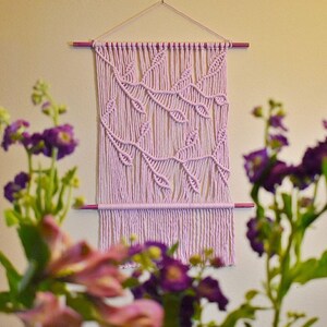 Puede incluir: Un tapiz de macramé de color lila claro con un patrón de hojas. El tapiz está hecho con cordón de algodón y tiene una varilla de madera en la parte superior e inferior.
