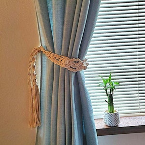 May include: A light blue curtain with a white macrame curtain tieback. The tieback has a tassel on each end. A small potted plant is visible in the background.