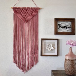 May include: A dusty rose macrame wall hanging with a triangular pattern and fringe. The hanging is displayed on a white wall with a wooden frame with the word "Thankful" inside, a framed picture with a white flower, and a vase with a pink flower.