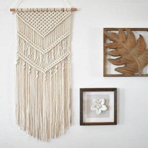 May include: A white macrame wall hanging with a geometric pattern and fringe. The hanging is displayed on a white wall with two framed pieces of art, one with a wooden leaf design and the other with a white flower.