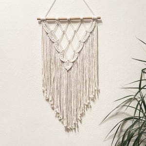 May include: A white macrame wall hanging with a geometric design and fringe. The hanging is attached to a wooden dowel rod.