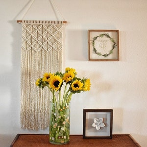 May include: A white macrame wall hanging with a geometric pattern, a vase of sunflowers, and a framed sign that says "enjoy life" in cursive.