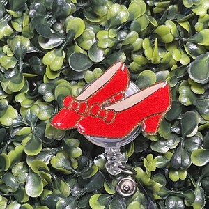 May include: A badge reel featuring a pair of bright red glittery shoes with a small bow on the front. The shoes have a tan interior lining and gold glitter trim along the edges. The badge reel mechanism is circular with a clear plastic cord.