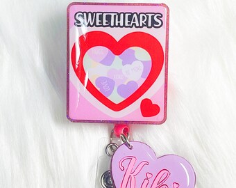 May include: A pink and glittery badge reel with a "Sweethearts" design. The design features a red heart with smaller purple hearts inside, each with a message. A lavender heart charm with the name "Kiki" hangs below.