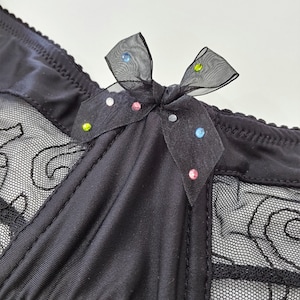 Puede incluir: Lencería negra con un panel de malla transparente y un lazo de satén negro con pedrería de colores.