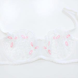 May include: White bra with floral lace detailing and pink accents. The bra features delicate lace cups with pink embroidered flower shapes. The straps are thin and white, and the overall design is feminine and delicate.