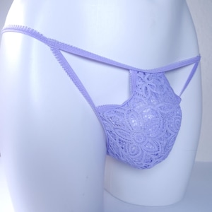 May include: A light purple lace thong featuring a floral pattern with thin, delicate side straps and a scalloped edge finish. The garment is constructed from a sheer, intricate lace fabric that forms a pouch-like front section.