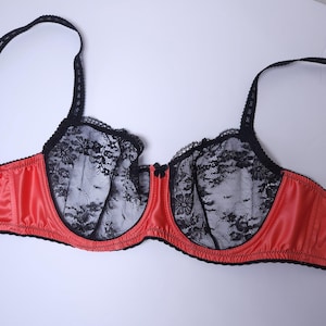 Puede incluir: Un sujetador rojo con detalles de encaje negro. El sujetador presenta un acabado satinado en las copas y una delicada superposición de encaje negro. Las correas y el ribete son negros, y un pequeño lazo acentúa el centro.