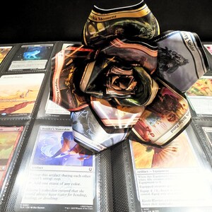 May include: A handmade paper flower made from Magic: The Gathering trading cards, resting on a binder page filled with various cards. The flower is composed of multiple card petals, showcasing vibrant colors and card art.