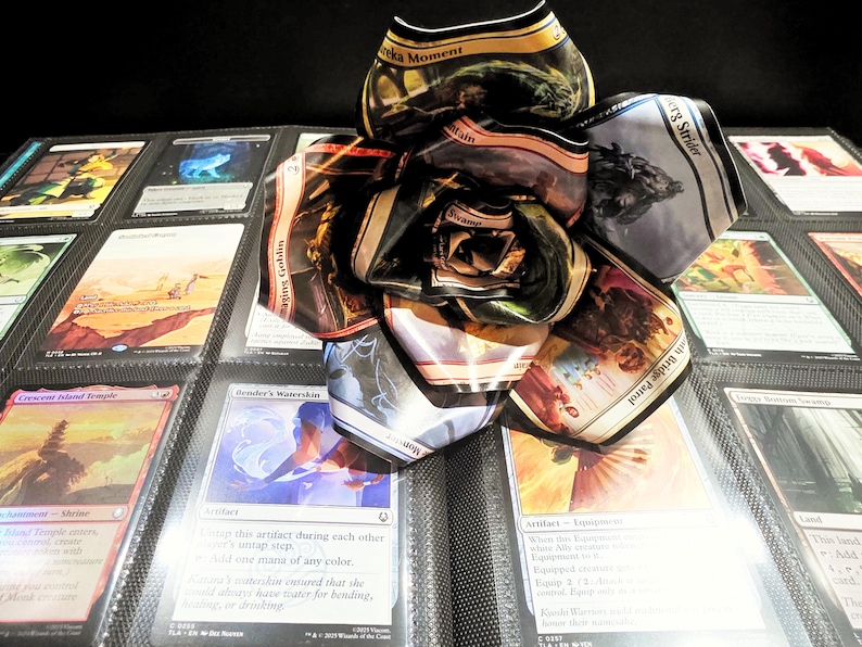 May include: A handmade paper flower made from Magic: The Gathering trading cards, resting on a binder page filled with various cards. The flower is composed of multiple card petals, showcasing vibrant colors and card art.