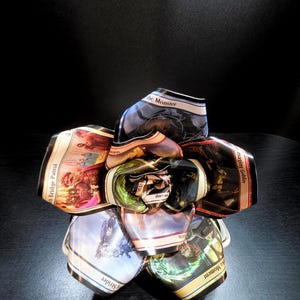 May include: A handcrafted, unique flower sculpture made from collectible trading cards. The petals display colorful fantasy art and text from the cards, arranged in a circular shape. The sculpture sits on a dark surface.