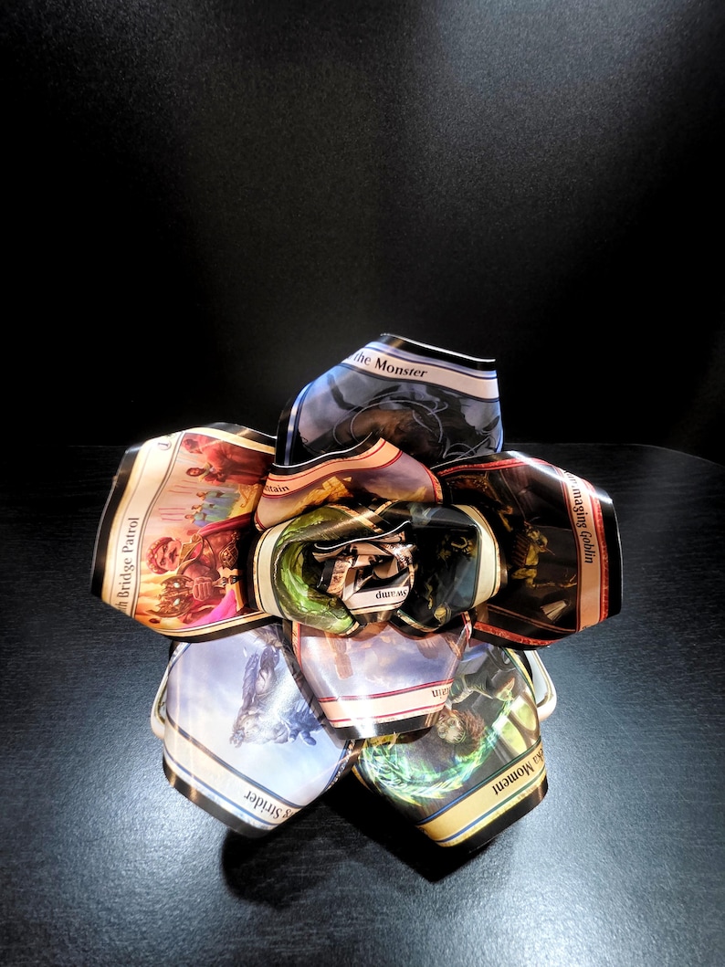 May include: A handcrafted, unique flower sculpture made from collectible trading cards. The petals display colorful fantasy art and text from the cards, arranged in a circular shape. The sculpture sits on a dark surface.