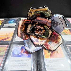 May include: A handmade paper flower made from Magic: The Gathering trading cards. The flower is in full bloom, with petals displaying various card art and text. The flower rests on a binder filled with more trading cards.