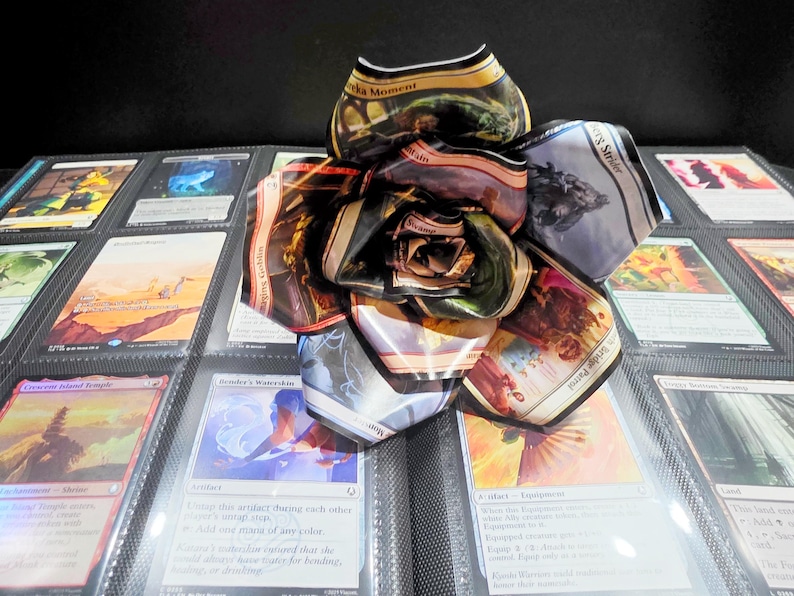 May include: A handmade paper flower made from Magic: The Gathering trading cards. The flower is in full bloom, with petals displaying various card art and text. The flower rests on a binder filled with more trading cards.