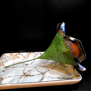 May include: A decorative serving tray with a white marble design and gold accents. A green leaf-shaped object rests on the tray, with a stem extending across the surface. A colorful label is partially visible.