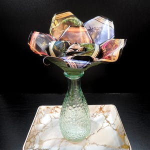 May include: A unique floral arrangement made from folded cards, displayed in a green glass vase. The vase sits on a rectangular tray with a marble design and gold accents. The background is black.