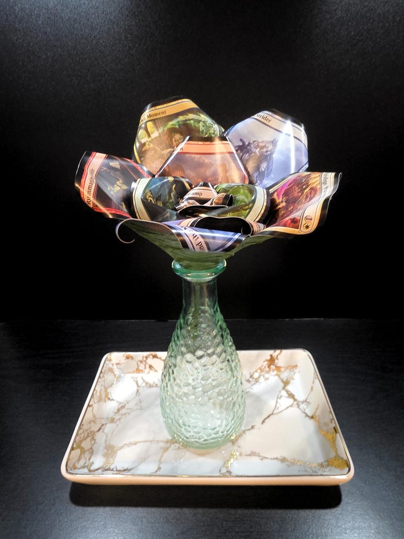 May include: A unique floral arrangement made from folded cards, displayed in a green glass vase. The vase sits on a rectangular tray with a marble design and gold accents. The background is black.